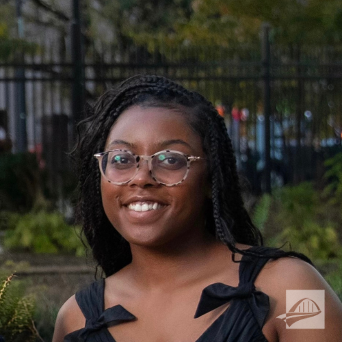 Amoirie Perteet headshot. Amoirie is wearing a black sleeveless top with bows on the straps. She is wearing tortoiseshell glasses. She has long black hair in small braids. She is outside with city park background that is blurred.