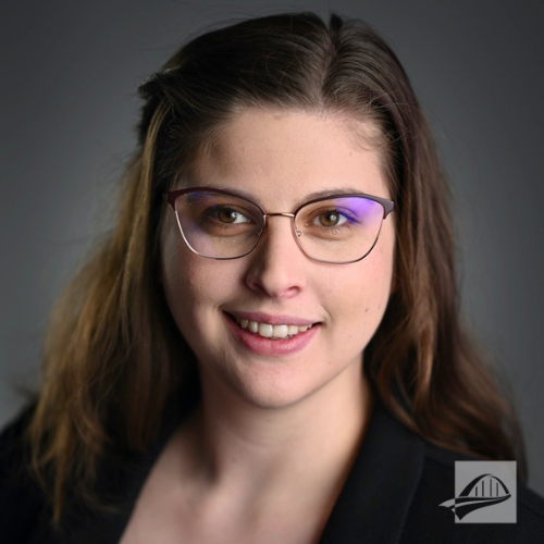 Emmalee berger headshot. Emmalee is wearing a black shirt a with a black cardigan over it. She has silver metal rimmed glasses and long brown hair.