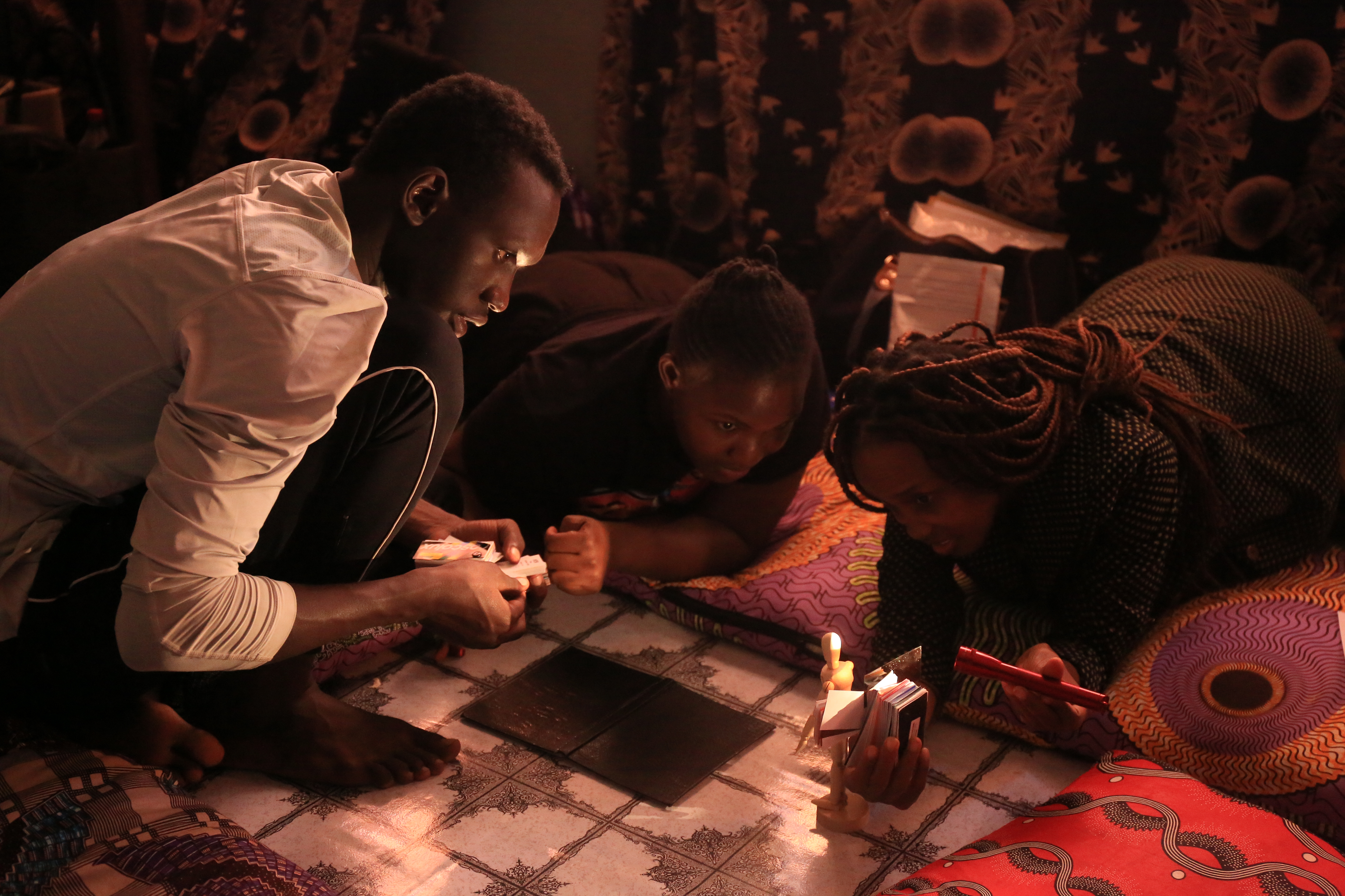 Two people lying on the floor and one squatting. One person in holing a light ad they're all looking at an object on the floor.