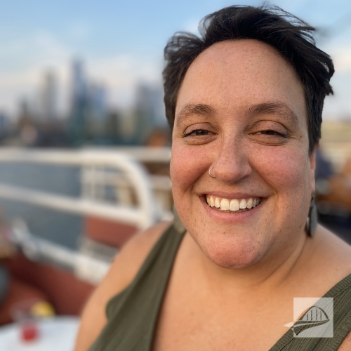 Nickie Dubick headshot. Nicki is wearing an olive green tank top, has short brown hair, a hoop nose ring, and large black circular earrings. She is standing outside with a blurred city skyline behind her.