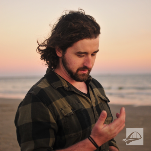 Zach Young headshot. Zach is wearing a green and black flannel buffalo check long sleeve shirt. He has medium length brown hair and is looking down at his right hand. He is wearing a brown hair tie around his wrist. He is walking on a beach at sunset and the ocean's shoreline and sand are both visible in the background.