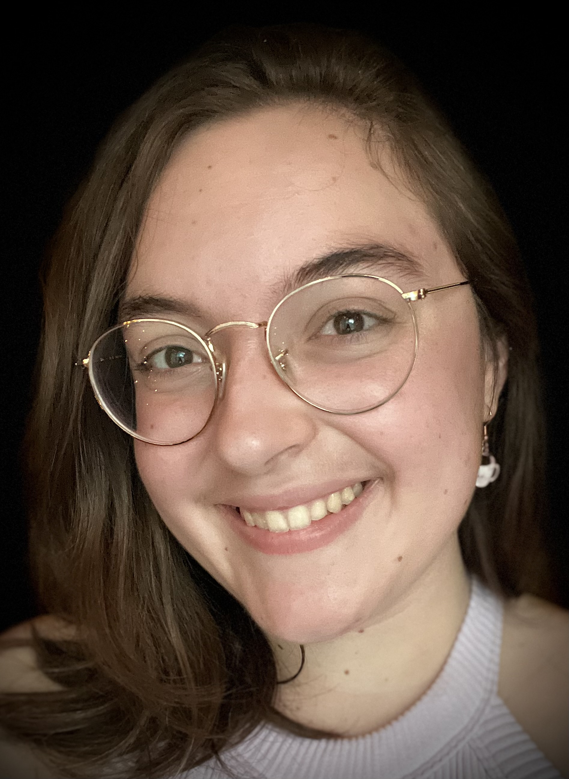 Abby Bilson headshot abby heas glasses brown hair and is smiling at the camera in a selfie-like style. She is wearing a white halter/tank top and teacup dangling earrings.