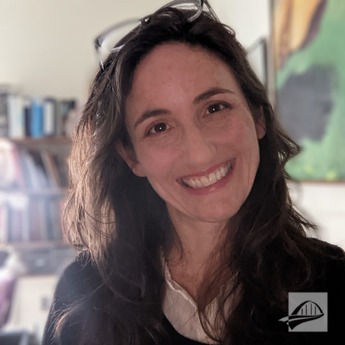 Charlene Gross headshot. Charlene is wearing a white button down collared shirt with a black sweater over it. She has long brown hair and is wearing a pair of eyeglasses on top of her head. She is tanding in an office with a bookshalef full of books behind her. A painting is behind her hung on the wall.