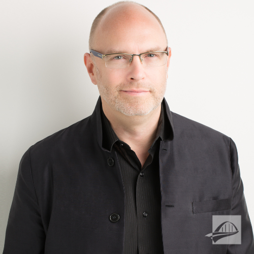 Clifton taylor headshot. Clifton is wearing an all black suit and black button down shirt. He does not have hair and is wearing glasses. He is standing against a white background