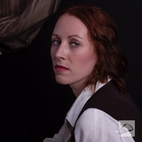 Daisy Long headshot. Daisy is wearing a longsleeve white button down collared shirt with a black vest over it. She has short red hair. She is sitting at an angle and is against a black background with a brown piece of fabric draped in the corner of the black background.