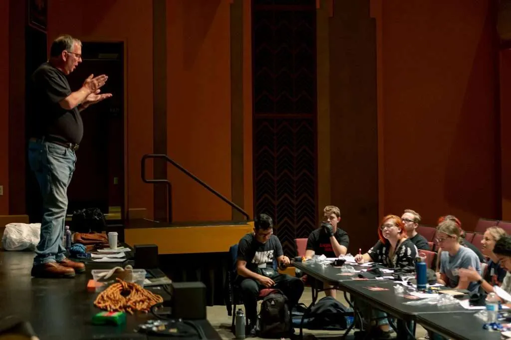 Dana Taylor standing on a stage in a black t shirt and jeans talking to a group of students sitting in the audience