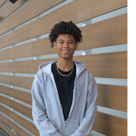 Darius Knight Evans is stading in front of a wood slatted wall and is wearing a black t shirt with a gray zip up hoodie over it and a thin gold chain. He is smiling at the camera and has black curly hair.