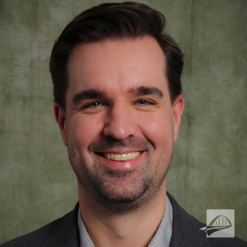 David Bisaha headshot. David is wearing a light blue button down shirt with a black suit coat over it. He has short brown hair. He is standing in front of a neutral cream background.