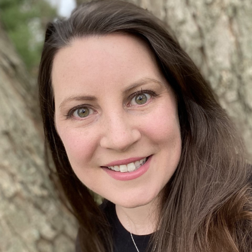 Dr Jennifer Leigh Sears Scheier headshot. She is wearing a black shirt with a silver necklace. She has long brown hair. She is standing outside in front of a tree.