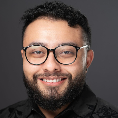 Frankie Charles headshot. Frankie is wearing a black floral button down shirt. He has a dark black beard and short black curly hair. He is wearing small black round stud earrings and black glasses.