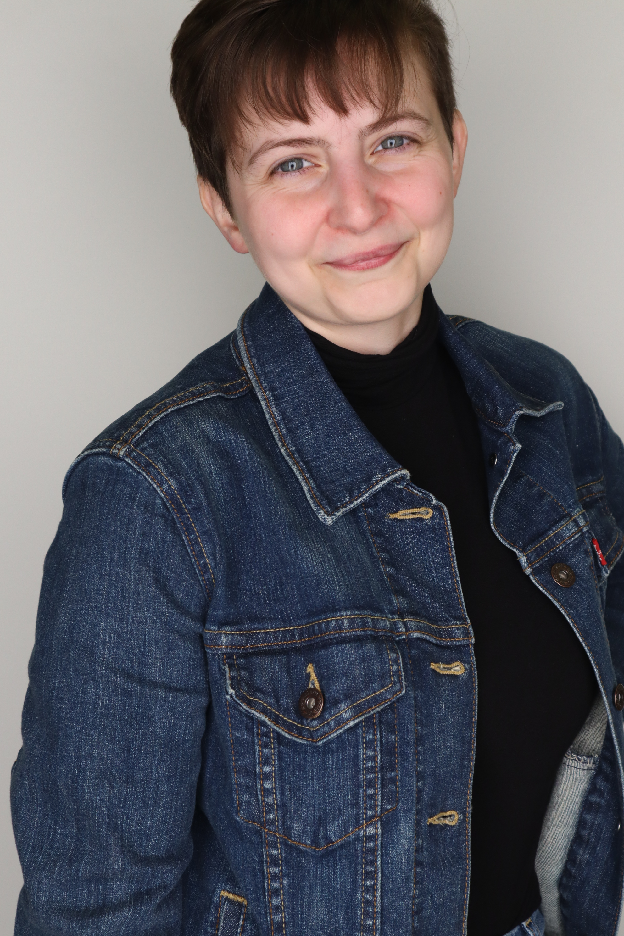 Natalie Houle Headshot. Natalie has short brown hair and is wearing a black turtle neck with a denim jacket over it. She is smiling directly into the camera in front of a gray/white background.