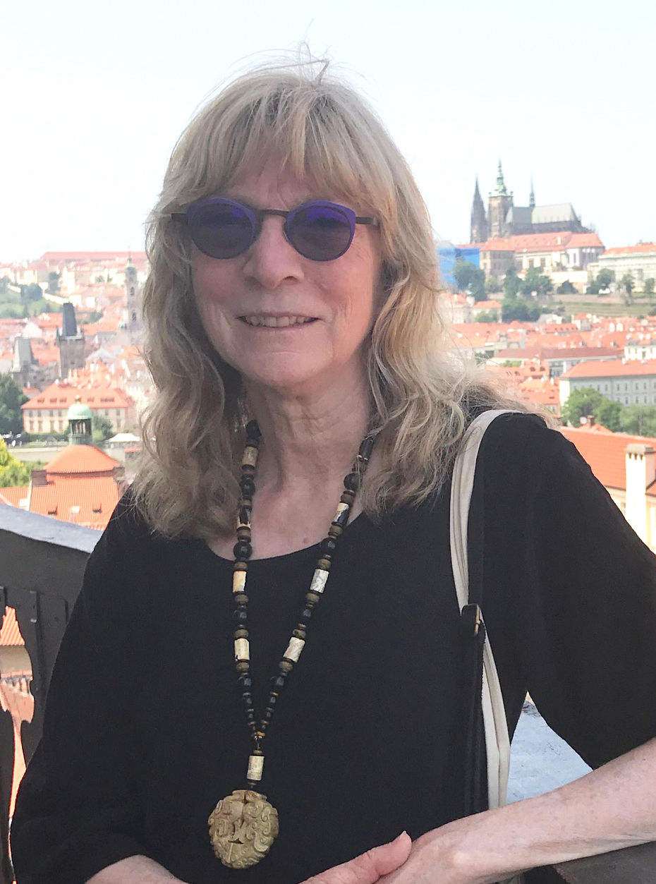 Sandy Bonds headshot. Sandy has blonde hair and is wearing a black tshirt with a tan and black necklace with a large stone hanging from it, and sunglasses. She is standing in front of a castle in Prague.